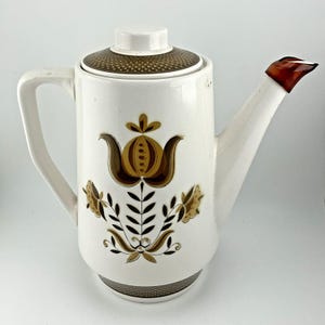 May include: White ceramic coffee pot with a brown floral design. The pot has a matching lid and a brown handle on the spout. The handle is white and the lid has a brown dotted pattern.