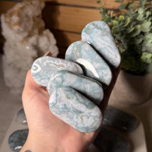 May include: A collection of polished, oval-shaped stones in shades of blue, green, and white. The stones have a marbled appearance and are held in a hand. The background includes a plant and other stones.