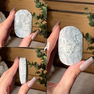 May include: A polished, oval-shaped stone with a white base and intricate green veining. The stone is held in a hand, showcasing its smooth surface and natural patterns. The stone's dimensions are approximately 3 inches long and 2 inches wide.