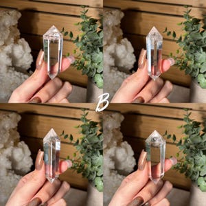 May include: Clear quartz crystal point, held against a backdrop of greenery and a wooden surface. The crystal is transparent with a pointed tip. The letter "B" is visible in the center of the image. The crystal is a popular item in the crystal healing and metaphysical community.