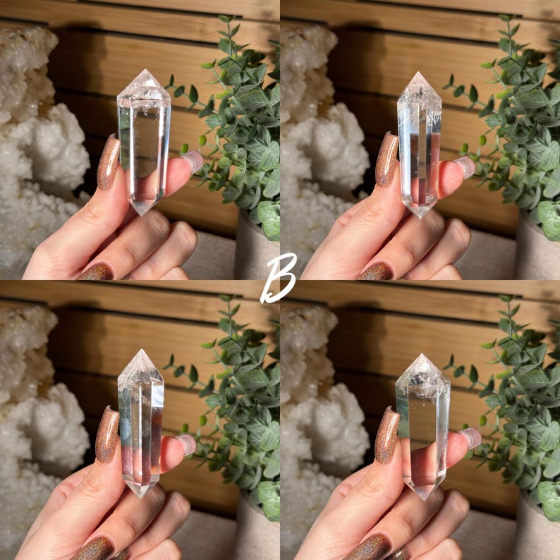 May include: Clear quartz crystal point, held against a backdrop of greenery and a wooden surface. The crystal is transparent with a pointed tip. The letter "B" is visible in the center of the image. The crystal is a popular item in the crystal healing and metaphysical community.