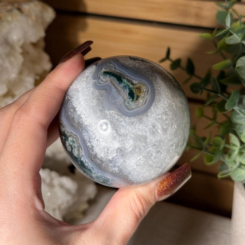 May include: A polished agate sphere with a mix of gray, white, and green patterns. The gemstone ball is held in a hand, showcasing its smooth, rounded shape. The sphere is a decorative item.