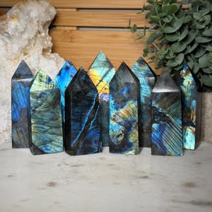 Puede incluir: Una colección de nueve torres de cristal de labradorita pulida dispuestas verticalmente. Cada piedra presenta una base gris oscuro con destellos iridiscentes de luz azul, dorada y amarilla sobre las superficies facetadas.