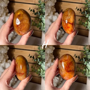 Puede incluir: Cuatro im&aacute;genes de una piedra preciosa pulida de forma ovalada. La piedra tiene un color &aacute;mbar c&aacute;lido con vetas de color marr&oacute;n rojizo. La piedra preciosa se sostiene en una mano, mostrando su superficie lisa y brillante y sus patrones naturales. El fondo incluye vegetaci&oacute;n y una superficie de madera.