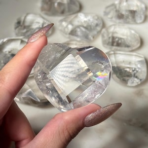 Puede incluir: Un coraz&oacute;n de cristal de cuarzo transparente con superficies facetadas, sostenido en una mano. El cristal en forma de coraz&oacute;n refleja la luz, creando un efecto arco&iacute;ris. Otros cristales en forma de coraz&oacute;n son visibles en el fondo.