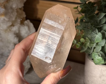 May include: A translucent, pale peach-colored quartz crystal point with a hexagonal shape and distinct horizontal striations along its faceted sides. The crystal exhibits natural internal inclusions and a smooth, polished surface texture throughout.