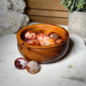 May include: A wooden bowl filled with polished agate stones in shades of red, orange, and white. Two additional stones rest outside the bowl. The bowl is made of a warm-toned wood, and the stones have a smooth, rounded shape.