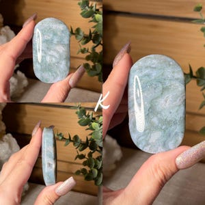 May include: A polished, oval-shaped stone with a light green and white marbled pattern. The stone has a smooth, rounded top and a flat bottom. The stone is held in a hand, showcasing its natural beauty and unique veining.