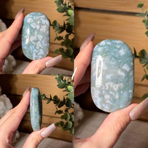 May include: A polished, oval-shaped gemstone with a mottled pattern of light blue and white. The stone is held in a hand, showcasing its smooth surface and natural variations. The gemstone is a decorative item.