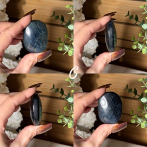 May include: Four images of a blue tiger eye stone, held in a hand. The stone is oval-shaped and polished, with visible chatoyancy. The stone's color ranges from dark blue to grey, with streaks of lighter hues. The background includes greenery and a wooden surface.