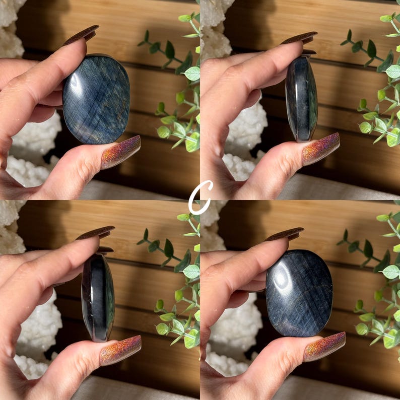 May include: Four images of a blue tiger eye stone, held in a hand. The stone is oval-shaped and polished, with visible chatoyancy. The stone's color ranges from dark blue to grey, with streaks of lighter hues. The background includes greenery and a wooden surface.