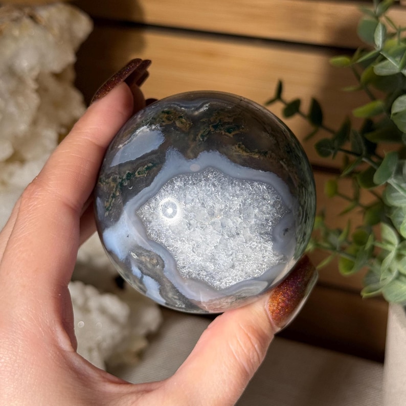 May include: A polished, spherical gemstone with a geode center. The orb features a mix of colors, including white, gray, brown, and green. The geode center has a crystalline appearance. The sphere is held in a hand.