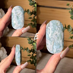 May include: A polished, oval-shaped stone with a marbled pattern of pale green, white, and gray. The stone is held in a hand, showcasing its smooth, rounded form. The natural stone has a unique, organic appearance.