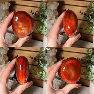 Puede incluir: Una piedra de cornalina pulida, de forma ovalada, que muestra una vibrante mezcla de tonos naranja y rojo. La superficie de la piedra es lisa y reflectante, con patrones naturales visibles. La piedra preciosa se sostiene en una mano, destacando su tama&ntilde;o y forma. Es un cristal natural.