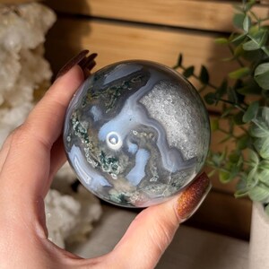 May include: A polished, spherical gemstone with a swirling pattern of gray, white, and green. The orb is held in a hand, showcasing its intricate natural design. The stone's surface reflects light, highlighting its unique mineral formations.
