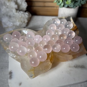May include: A collection of rose quartz spheres displayed in a decorative, heart-shaped dish. The spheres vary in shades of pink and are arranged to fill the dish. The dish is a light, translucent color, resting on a white surface.