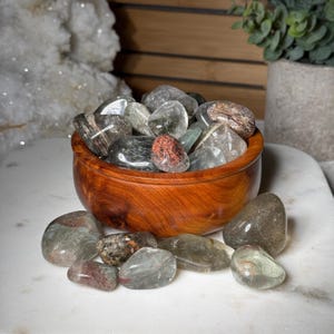 Puede incluir: Un cuenco de madera lleno de piedras pulidas, translúcidas, verdes y grises. Algunas piedras están esparcidas alrededor del cuenco sobre una superficie blanca. El cuenco es de madera marrón rojiza.