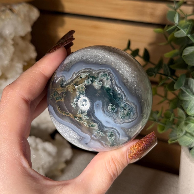 May include: A polished, spherical gemstone with a swirling pattern of gray, white, green, and brown. The orb is held in a hand, showcasing its intricate natural design. The gemstone is a decorative item.