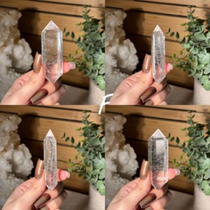 May include: Four clear quartz crystal points, each held in a hand. The crystals are transparent with visible internal structures. The background includes greenery and a wooden surface, suggesting a natural or spiritual theme.