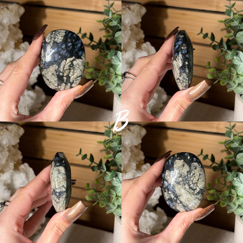May include: An oval-shaped stone with a polished surface. The stone features a mix of dark blue, gray, and white patterns. The stone is held in a hand, with the letter "B" visible in the background.