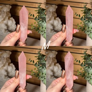 May include: Four rose quartz crystal obelisks, each held by a hand. The crystals are a soft pink color, with a polished, smooth surface. The obelisks are displayed against a backdrop of greenery and a wooden surface.