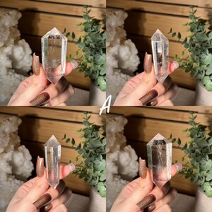 May include: Four images of a clear quartz crystal point. The crystal is held in a hand with long, polished nails. The crystal is transparent with visible internal structures. The letter "A" is visible in white text.