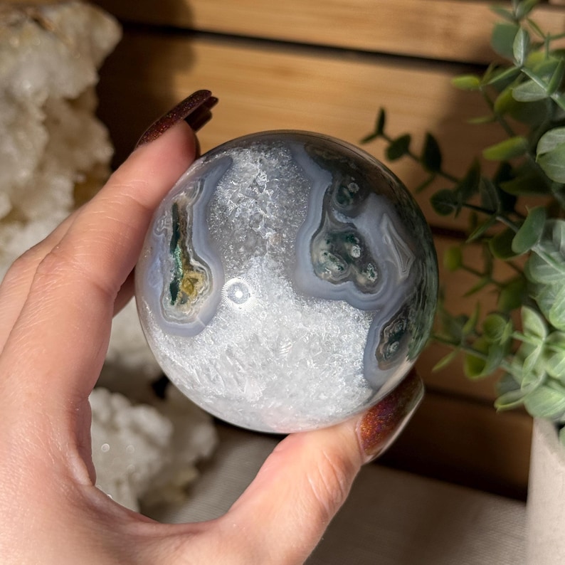 May include: A polished, spherical gemstone with a mix of white, gray, and green patterns. The orb is held in a hand, showcasing its natural textures and colors. The stone appears to be a decorative item or a collectible.