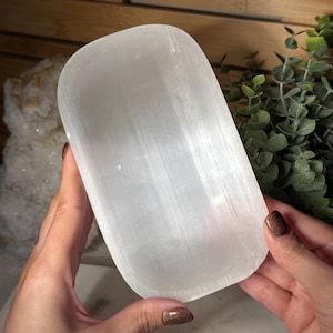 May include: A rectangular selenite charging plate with rounded corners, featuring a smooth, translucent white surface with a fibrous, pearlescent texture that reflects light across its polished, slightly concave interior surface.