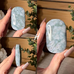 May include: A polished, oval-shaped stone with a swirling pattern of pale green and white. The stone has a smooth, rounded top and a flat bottom. The stone is held in a hand, showcasing its natural beauty and unique design.