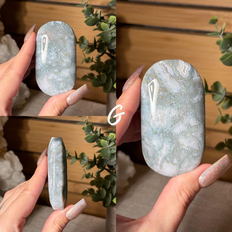 May include: A polished, oval-shaped gemstone with a swirling pattern of white and pale green. The stone is held up against a wooden background, showcasing its smooth, rounded form. The letter "G" is visible in the image.