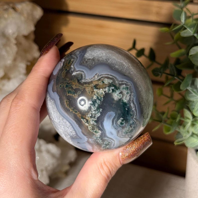 May include: A polished, spherical gemstone with a moss agate pattern. The orb features swirling patterns of gray, white, and green, creating a natural, organic aesthetic. The sphere is held in a hand, showcasing its size and detail.
