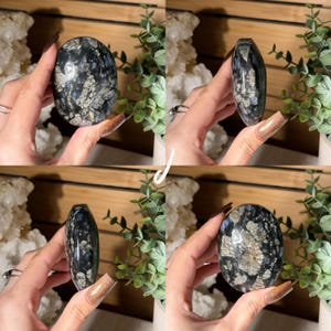 May include: A polished, oval-shaped gemstone with a dark base color, featuring intricate patterns of blue, gray, and white. The stone is held in a hand, showcasing its smooth, reflective surface. The gemstone is approximately 3 inches in length.