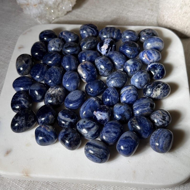 K&ouml;nnte beinhalten: Eine Sammlung polierter Sodalith-Steine, jeder in tiefblau mit wei&szlig;en Maserungen. Die oval geformten Steine liegen auf einem wei&szlig;en, quadratischen Teller. Die Natursteine variieren in Gr&ouml;&szlig;e und Muster und bilden eine einzigartige Pr&auml;sentation.