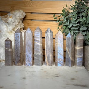 Puede incluir: Ocho torres de piedra pulida dispuestas en fila, cada una con forma de prisma hexagonal y punta. Las piedras presentan una superficie lisa de color gris parduzco con destellos azules iridiscentes y sutiles bandas horizontales.