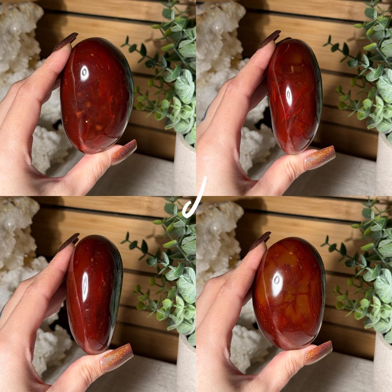 Puede incluir: Una piedra de jaspe rojo pulida, de forma ovalada. La piedra tiene una superficie lisa y brillante con variaciones naturales de color y patr&oacute;n. El jaspe se sostiene sobre un fondo de vegetaci&oacute;n y cristales blancos.
