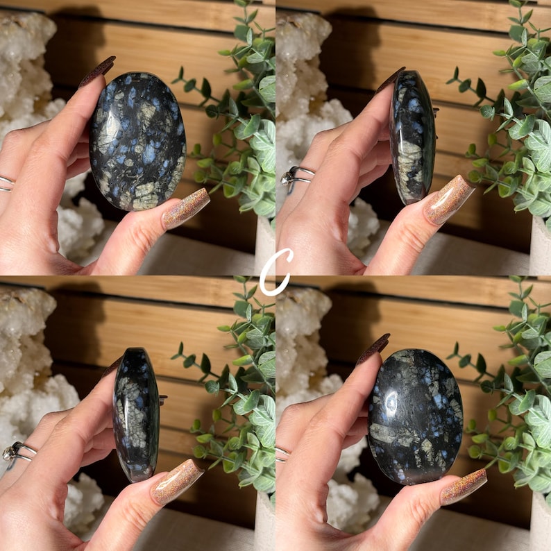 May include: A polished, oval-shaped stone with a dark base color, speckled with blue and gray patterns. The stone is held in a hand, showcasing its smooth, reflective surface. The stone's unique markings create a natural, artistic appearance.