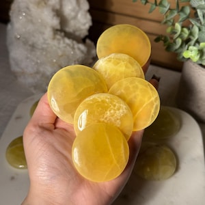 May include: A hand holds several polished, round yellow calcite stones. The stones vary in size and have a translucent appearance. The background includes a white surface and a potted plant, suggesting a natural or decorative theme.