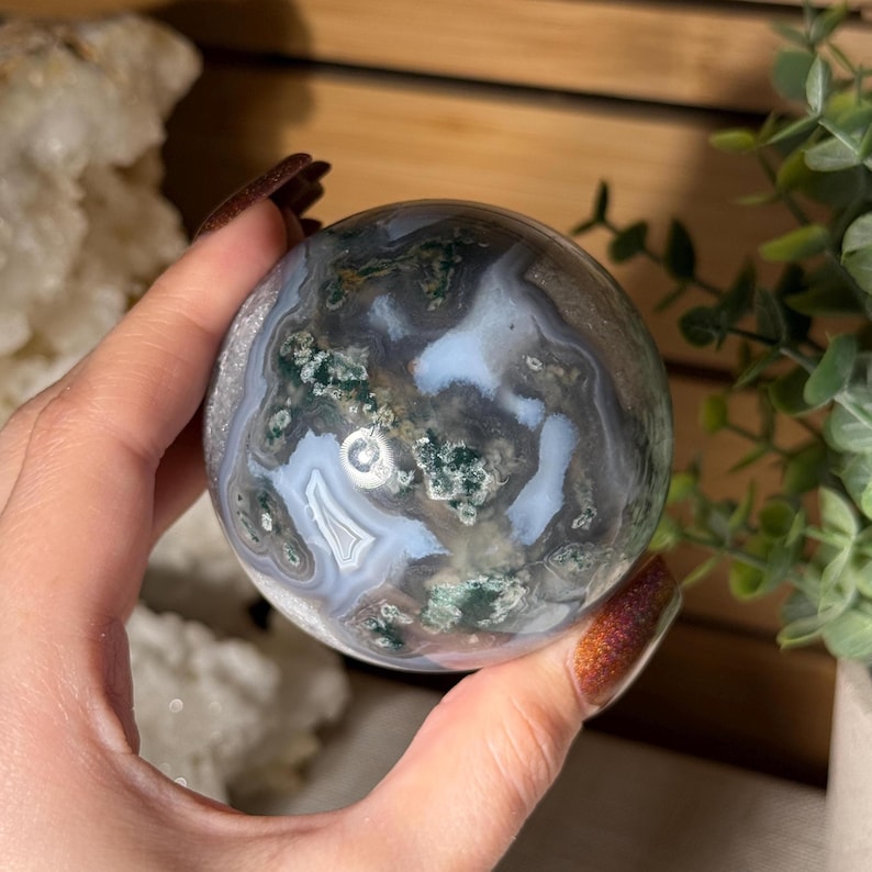 May include: A polished moss agate sphere, showcasing a blend of gray, white, and green patterns. The gemstone is spherical, with a smooth surface. The agate's natural colors and textures are visible, creating a unique decorative item.