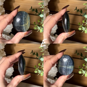 May include: Four views of a polished blue tiger's eye stone. The stone has a dark blue base color with golden-yellow and green chatoyancy. The stone is oval-shaped and held in a hand.