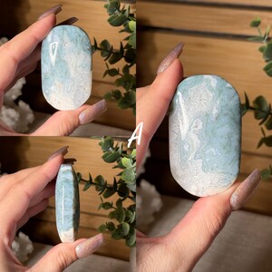 May include: A collection of polished, oval-shaped gemstones. The stones feature a blend of light blue and white patterns, with a smooth, glossy finish. The gemstones are displayed against a backdrop of greenery and wood.