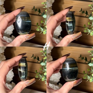 May include: An oval-shaped, polished gemstone with bands of dark blue, gold, and teal. The stone is held in a hand, showcasing its smooth, reflective surface. The gemstone is a natural, decorative item.