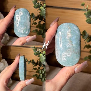 May include: A polished, oval-shaped gemstone with a light blue and white marbled pattern. The stone is held in a hand, showcasing its smooth surface and natural variations. The letter "H" is written on the image.