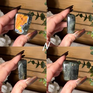 May include: Four images of polished labradorite crystals. One crystal has a vibrant orange and blue flash. The other three crystals are a mix of gray and green. The crystals are held up against a background of greenery.
