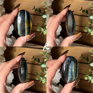 May include: Four images of a polished, oval-shaped gemstone. The stone has a dark blue base with streaks of gold and lighter blue. The gemstone is held in a hand, showcasing its smooth, reflective surface. The background includes greenery and white crystals.