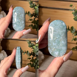 May include: A polished, oval-shaped gemstone with a mottled pattern of pale green, white, and gold. The stone is held in a hand, showcasing its smooth surface and natural variations. The gemstone is a decorative item.