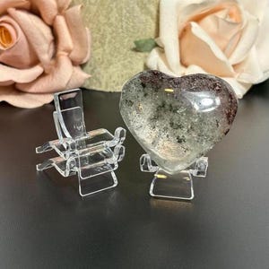 May include: A heart-shaped crystal with green and brown inclusions, displayed on a clear acrylic stand. The crystal is set against a backdrop of soft pink and cream-colored roses. A second clear acrylic stand is also visible.
