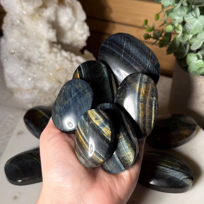 May include: A collection of polished blue tiger eye stones. The stones have a smooth, oval shape with streaks of gold and black. The stones are held in a hand, with several more stones visible in the background.