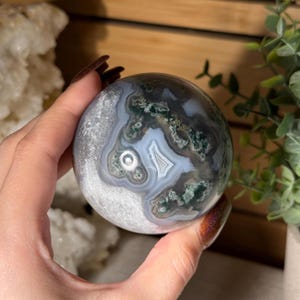 May include: A polished, spherical gemstone with a unique pattern of gray, white, and green swirls. The orb is held in a hand, showcasing its intricate natural design. The stone's surface has a smooth, reflective quality, highlighting its natural beauty.