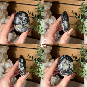 May include: A polished, oval-shaped stone with a dark blue base and a pattern of light gray and white inclusions. The stone is held in a hand, showcasing its smooth surface and natural variations. The stone is a unique, decorative item.