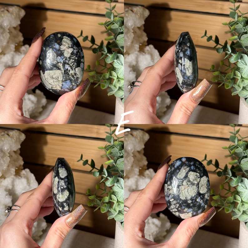 May include: A polished, oval-shaped stone with a dark blue base and a pattern of light gray and white inclusions. The stone is held in a hand, showcasing its smooth surface and natural variations. The stone is a unique, decorative item.